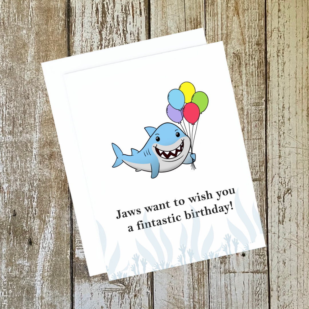 A photo of a birthday card. It has a shark on holding balloons. Text reads, Jaws want to wish you a fintastic birthday!