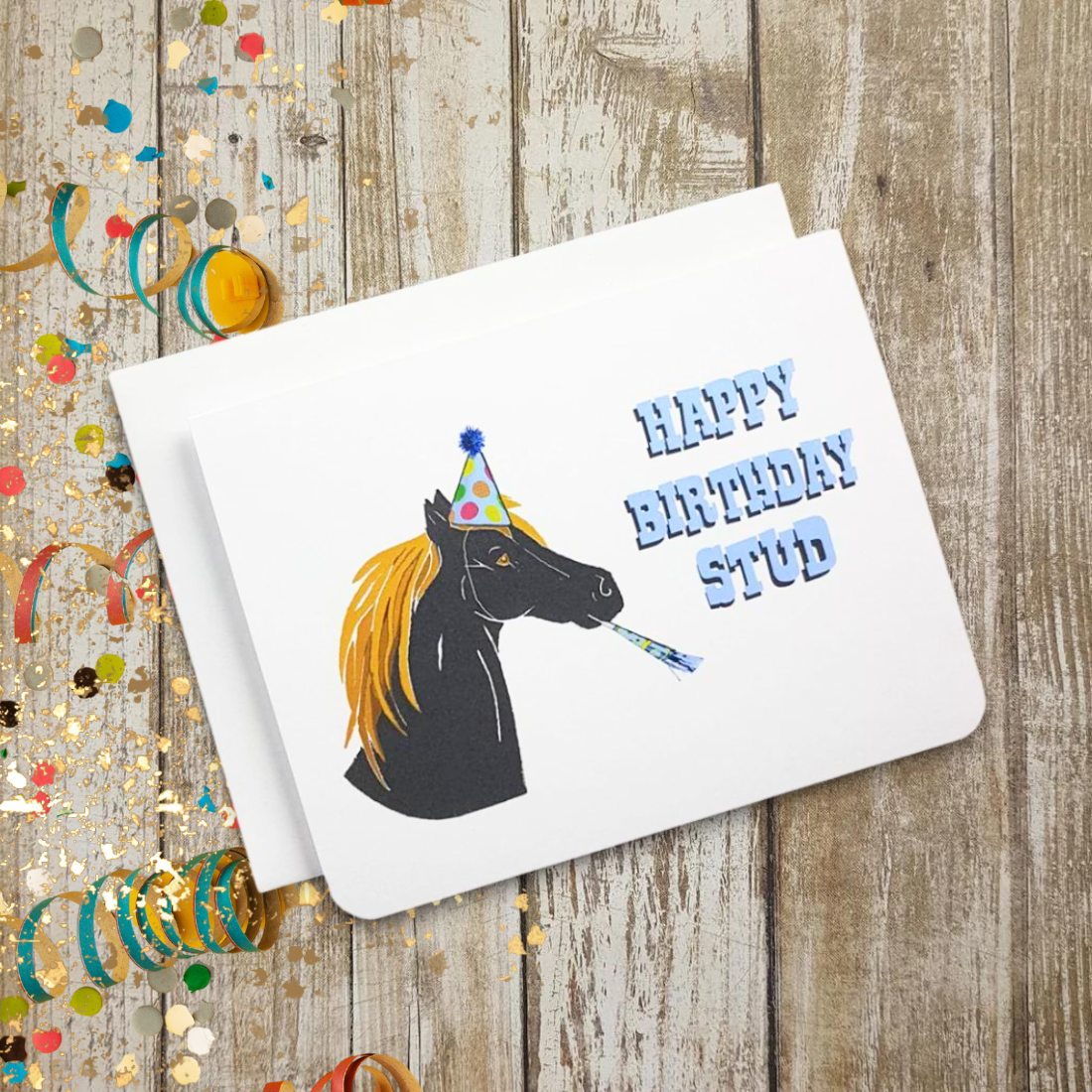 A photo of a white birthday card. It has an illustration of a black horse on it. It is wearing a party hat and has a streamer in its mouth. Blue text on card reads, 'Happy Birthday Stud.'