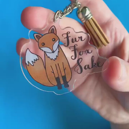A video of a hand showing of a key chain. The keychain has a cartoon fox on it. Text on keychain reads 'Fur Fox Sake.'