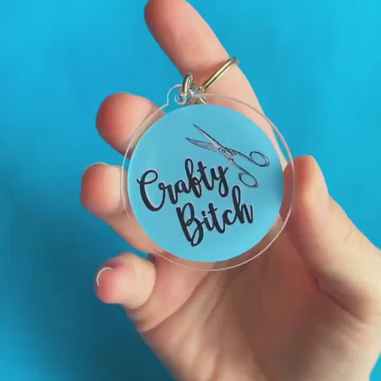 A video of someone showing off a key chain. It's round and blue. On it, there's pair of scissors. Text on keychain reads 'Crafty Bitch.'
