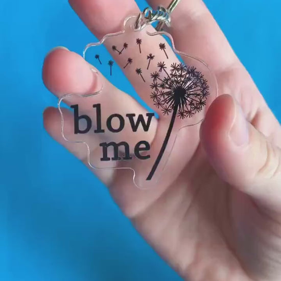 A video of a hand showing off a key chain. It has a blown dandelion on it. Text on key chain reads 'Blow Me.'