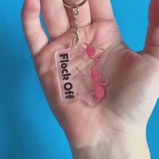 A video of a hand displaying a key chain. It has three flamingoes on it. Text below flamingos reads "Flock Off.'