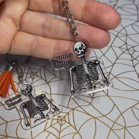 A video of a halloween keychain. The keychain has a skeleton on it. Text on keychain reads 'Wanna Bone?'